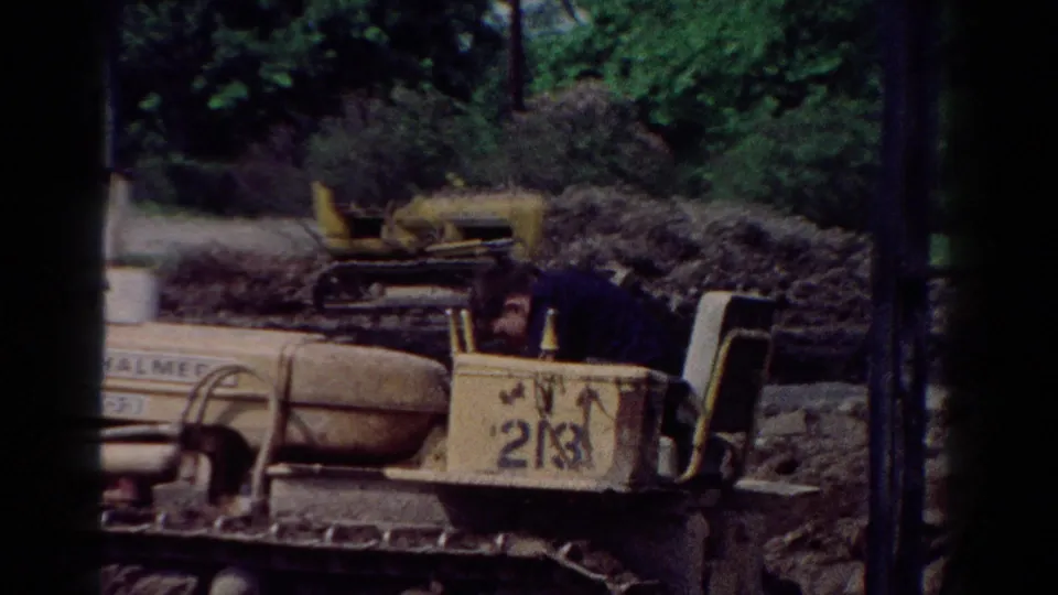 1954 Toledo home-movie frame showing a person seated on a yellow tracked bulldozer near piles of soil or brush.