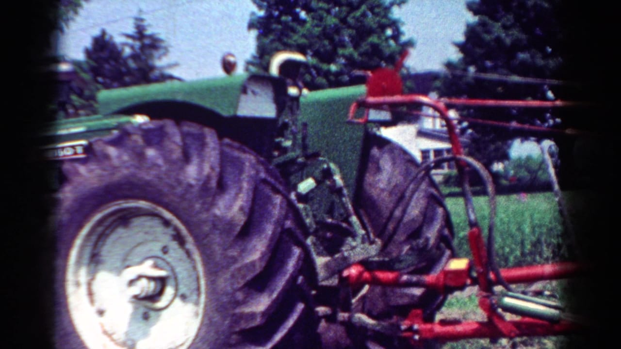 1954 Toledo-area home-movie frame showing a green tractor's rear tire and red attached farm implement near trees and grass.