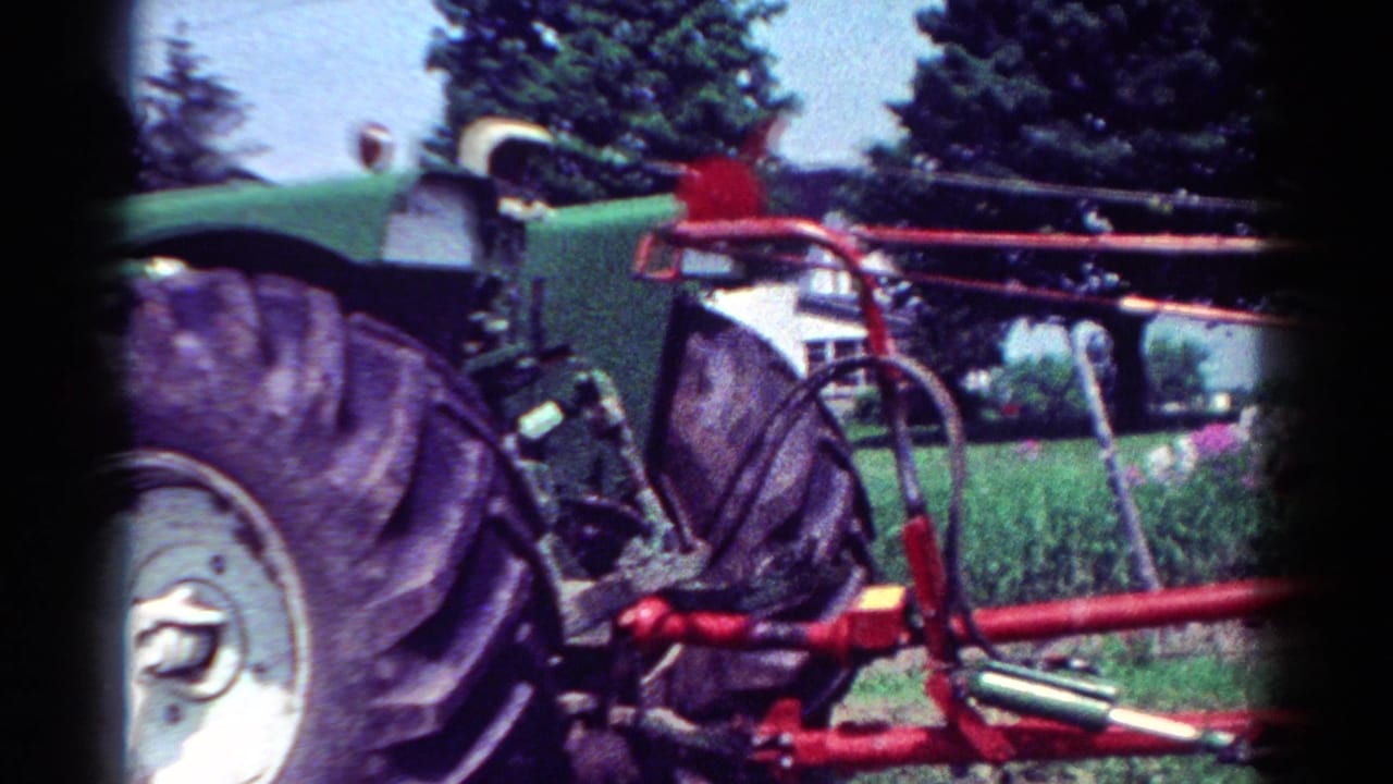 Green tractor and red farm implement seen from the side near trees.