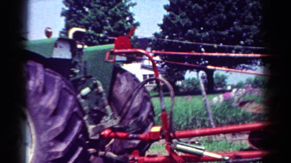 Home-movie frame from the 1954 Toledo clip showing tractor wheel detail and a red farm attachment against greenery.