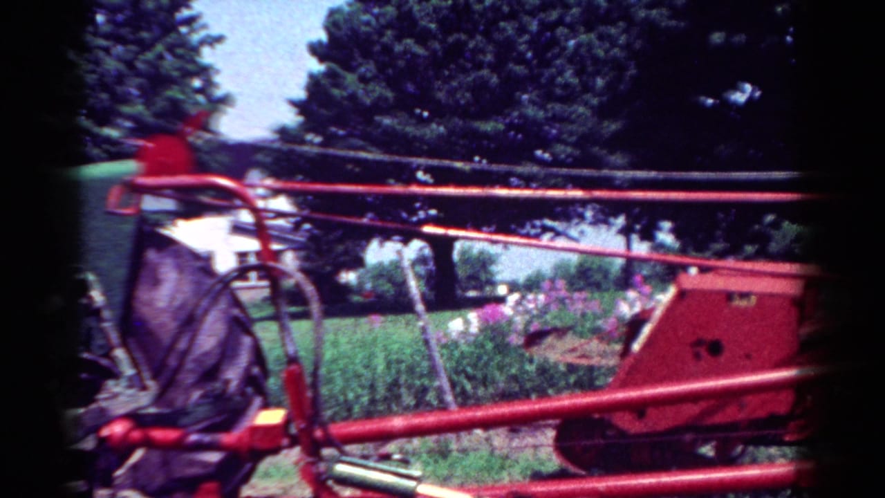 1954 Toledo home-movie frame showing a red tractor frame in front of lawn, flowers, trees, and a partial house view.