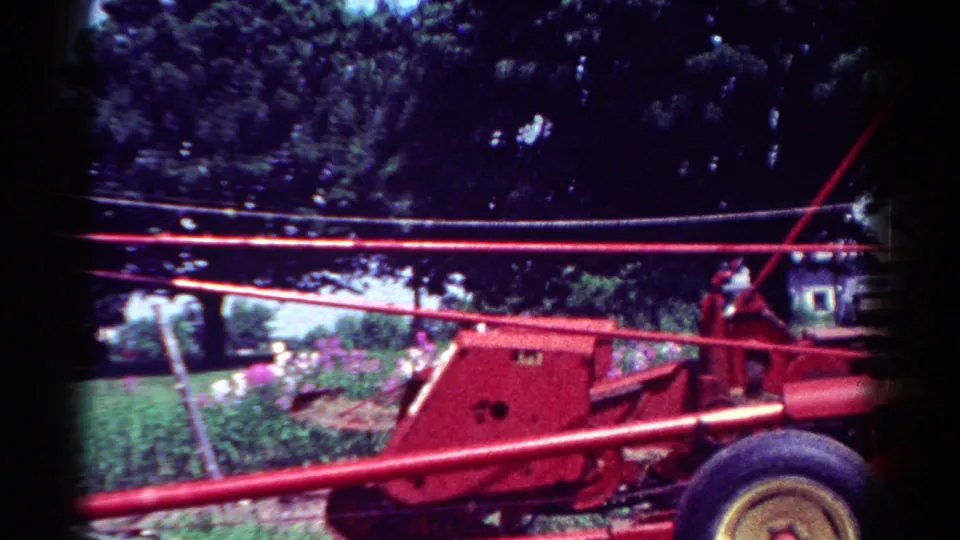 1954 Toledo home-movie still showing a red tractor body and wheel near a lawn, garden flowers, trees, and a distant house.