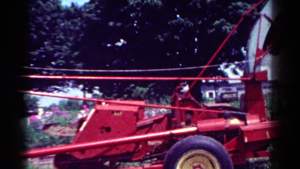 1954 Toledo 8mm frame showing the red tractor's wheel and frame beside a green yard, flower beds, dark trees, and a partial house.