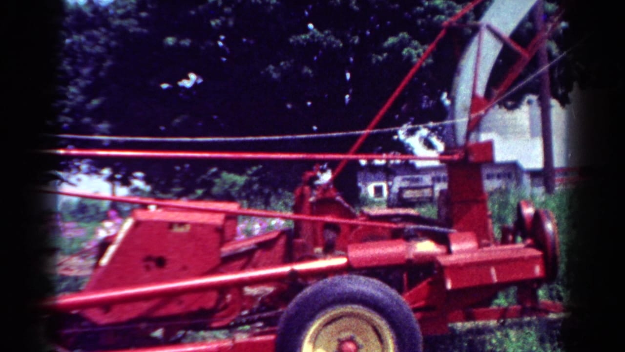 1954 Toledo home-movie frame showing a red wood chipper with a curved chute, wheel, trees, grass, and a house in the background.