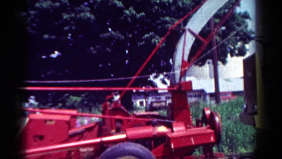 Red tow-behind chipper in a Toledo 1954 home-movie frame, with its raised chute, wheel, trees, grass, and house visible.