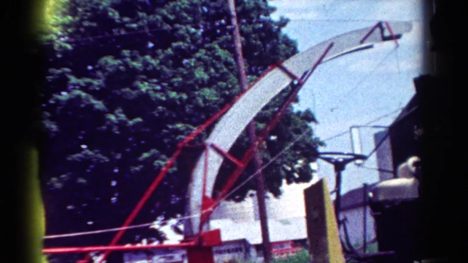 The chipper's curved discharge chute rises above the yard, with trees, overhead wires, a house, and part of a green dump truck visible.