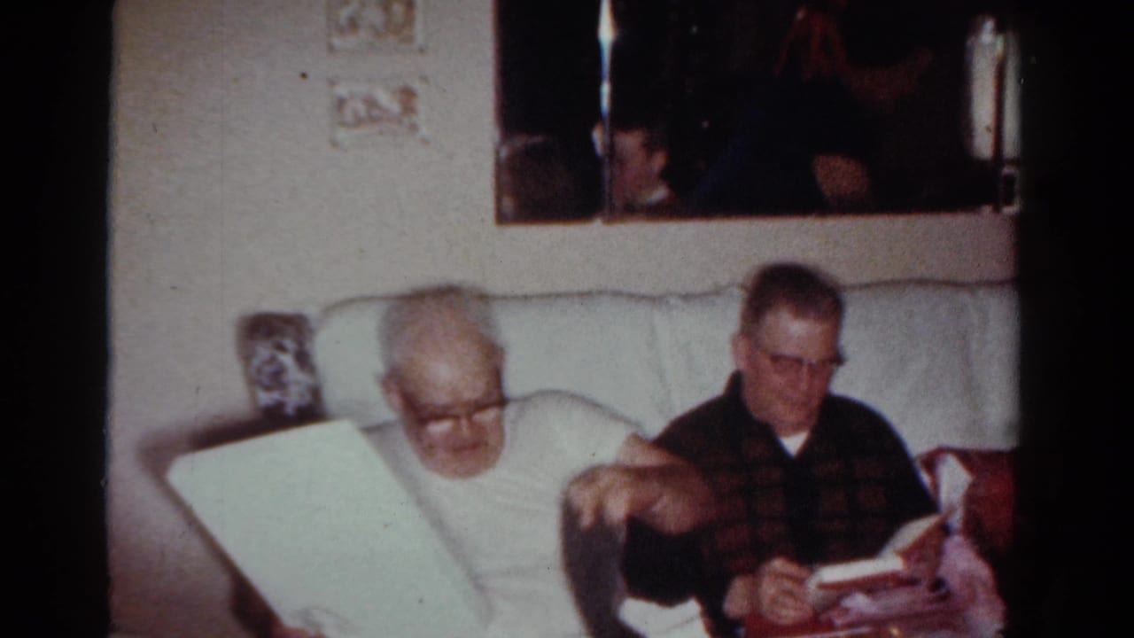 In 1954 Toledo home-movie footage, two men sit on a pale couch as one man in glasses looks down at a large wrapped item or book.