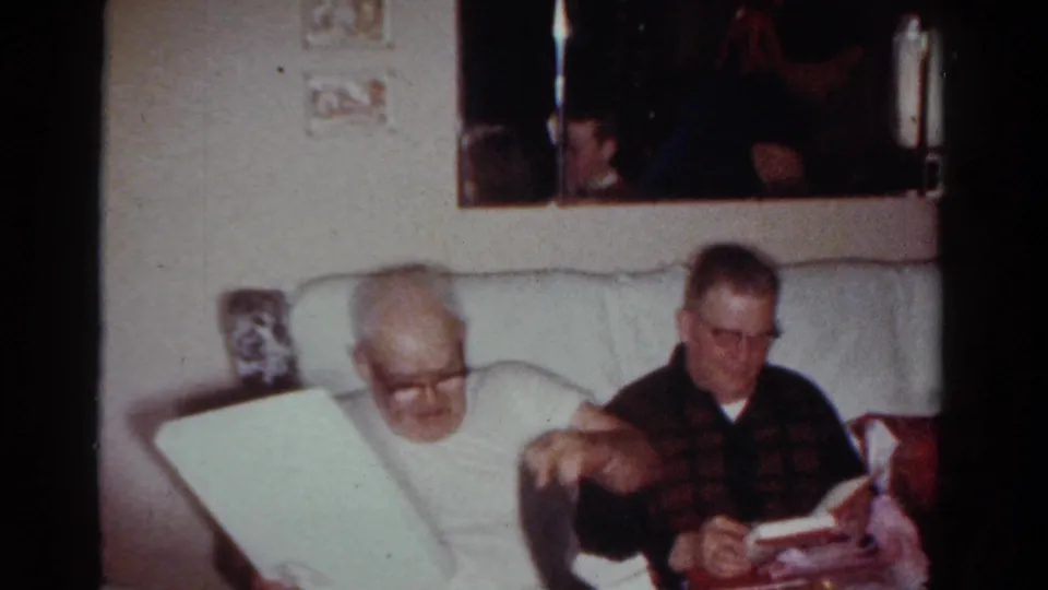 In 1954 Toledo home-movie footage, two men sit on a pale couch as one man in glasses looks down at a large wrapped item or book.