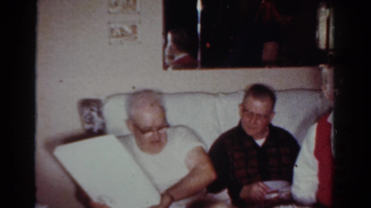 Two men seated in a Toledo living room look toward an opened gift or book, with a patterned sweater and pale couch visible.