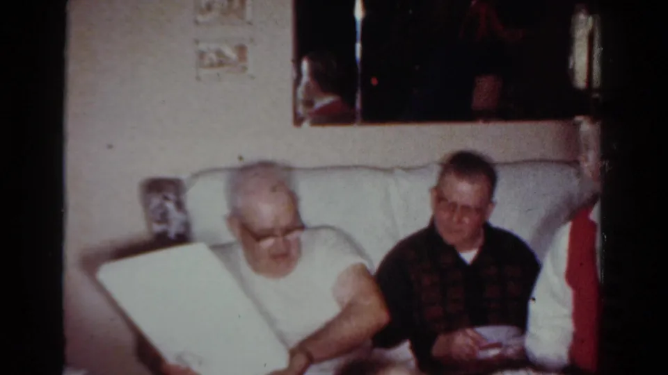 Two men seated in a Toledo living room look toward an opened gift or book, with a patterned sweater and pale couch visible.