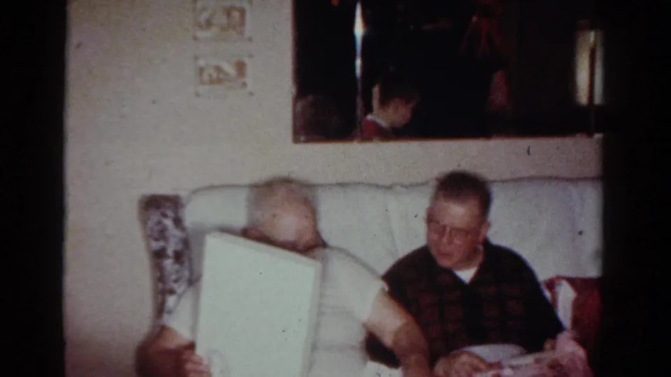 A man in a white shirt and glasses holds a large white object while another seated man looks on in the living room.