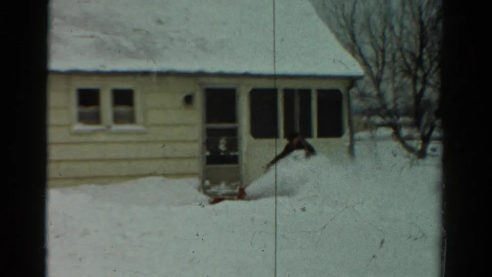 In 1954 Toledo, Ohio footage, a man in dark winter clothing clears deep snow near a light-colored house with a porch.