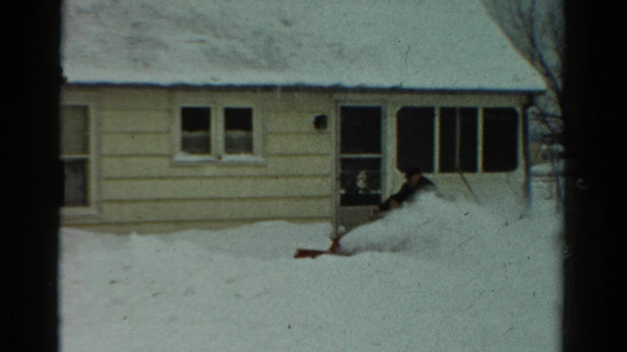 Man pushes a snow blower as snow sprays across the yard near the house.