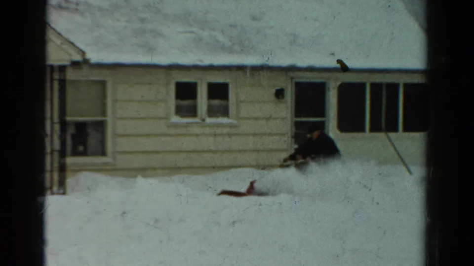 A man guides the snow blower through packed yard snow, with the house facade and enclosed porch visible behind him.
