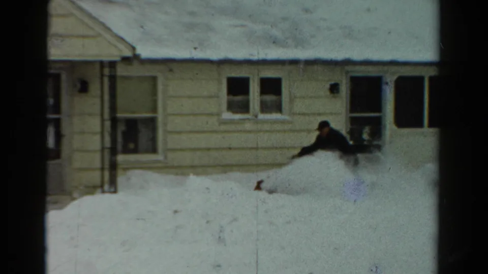 The 1954 Toledo home movie shows the man partly behind blowing snow while clearing near the house and porch windows.
