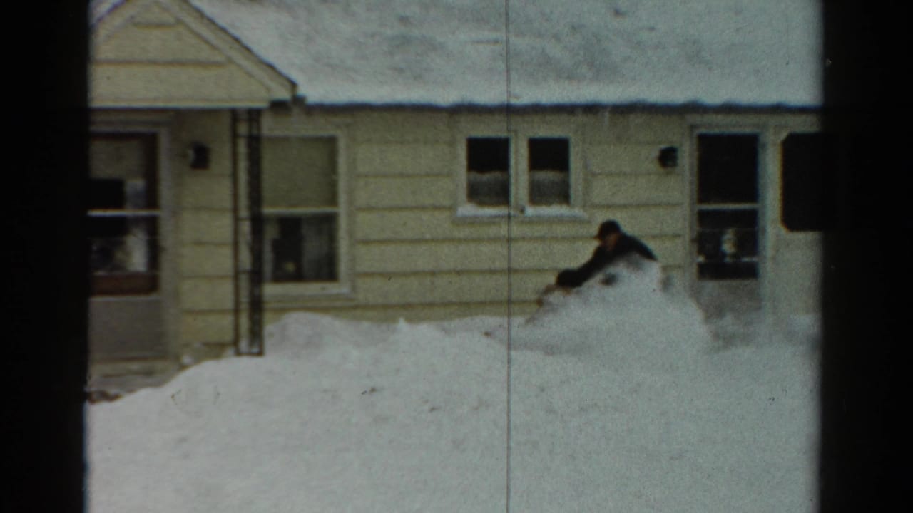 1954 Toledo home-movie frame of a man in dark winter clothing using a snow blower beside a yellow-sided house in deep snow.