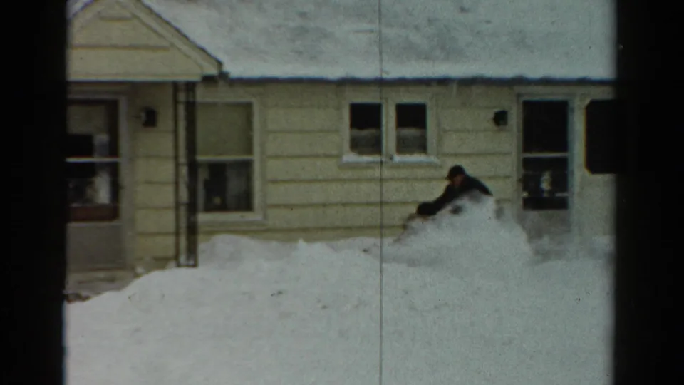 1954 Toledo home-movie frame of a man in dark winter clothing using a snow blower beside a yellow-sided house in deep snow.