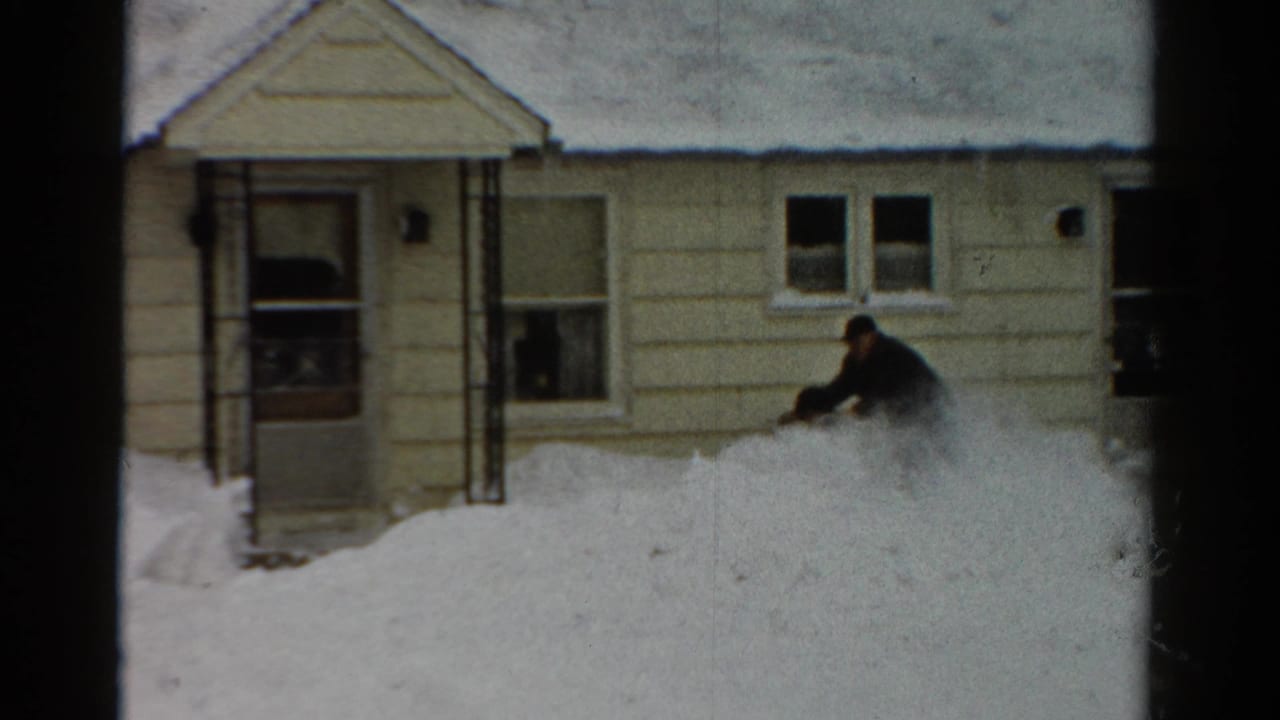 Man guiding a snow blower along a high snowbank in front of a house.