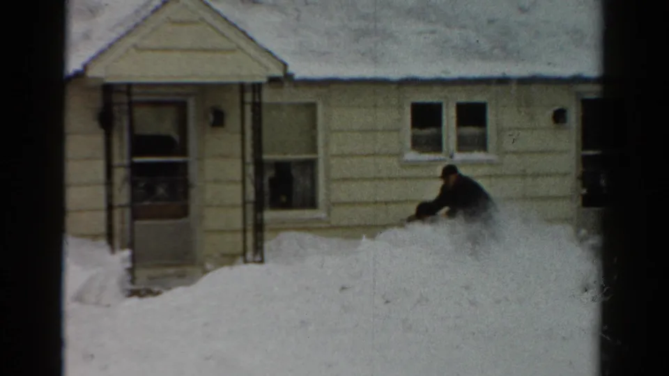 1954 Toledo frame showing a man working a snow blower through heavy snow near the front entrance of a residential house.
