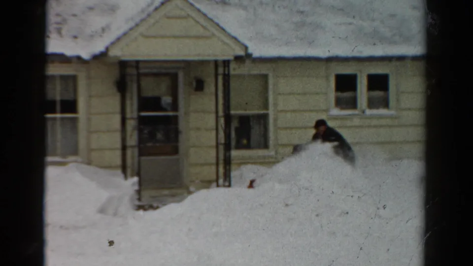1954 Toledo home-movie frame with the operator partly hidden behind a deep drift outside a snow-covered residential facade.