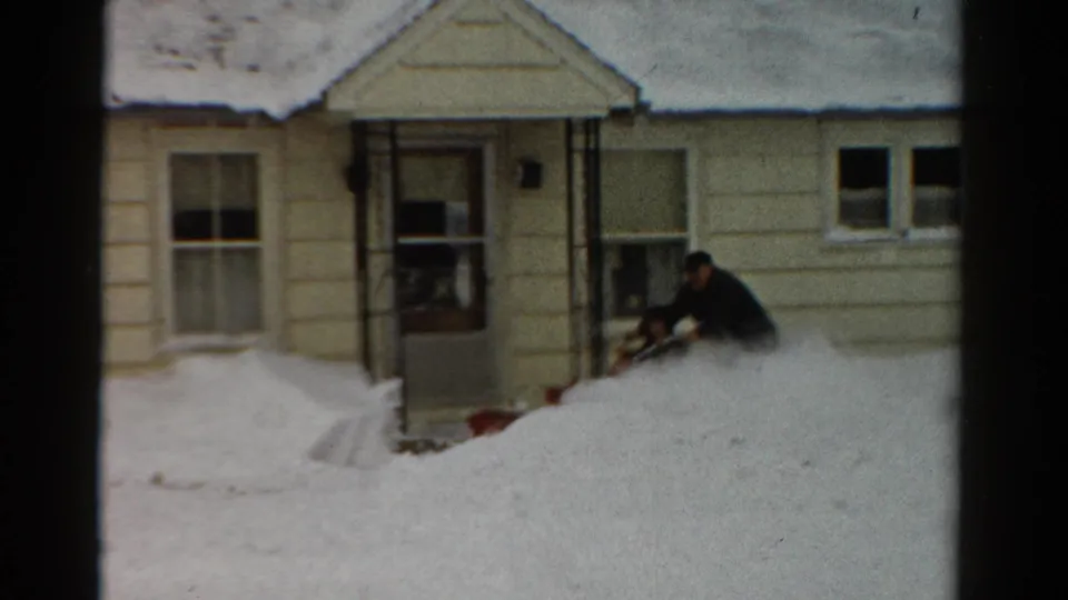 1954 Toledo frame of a man leaning into the snow blower as he clears a deep drift outside a yellow-sided home.