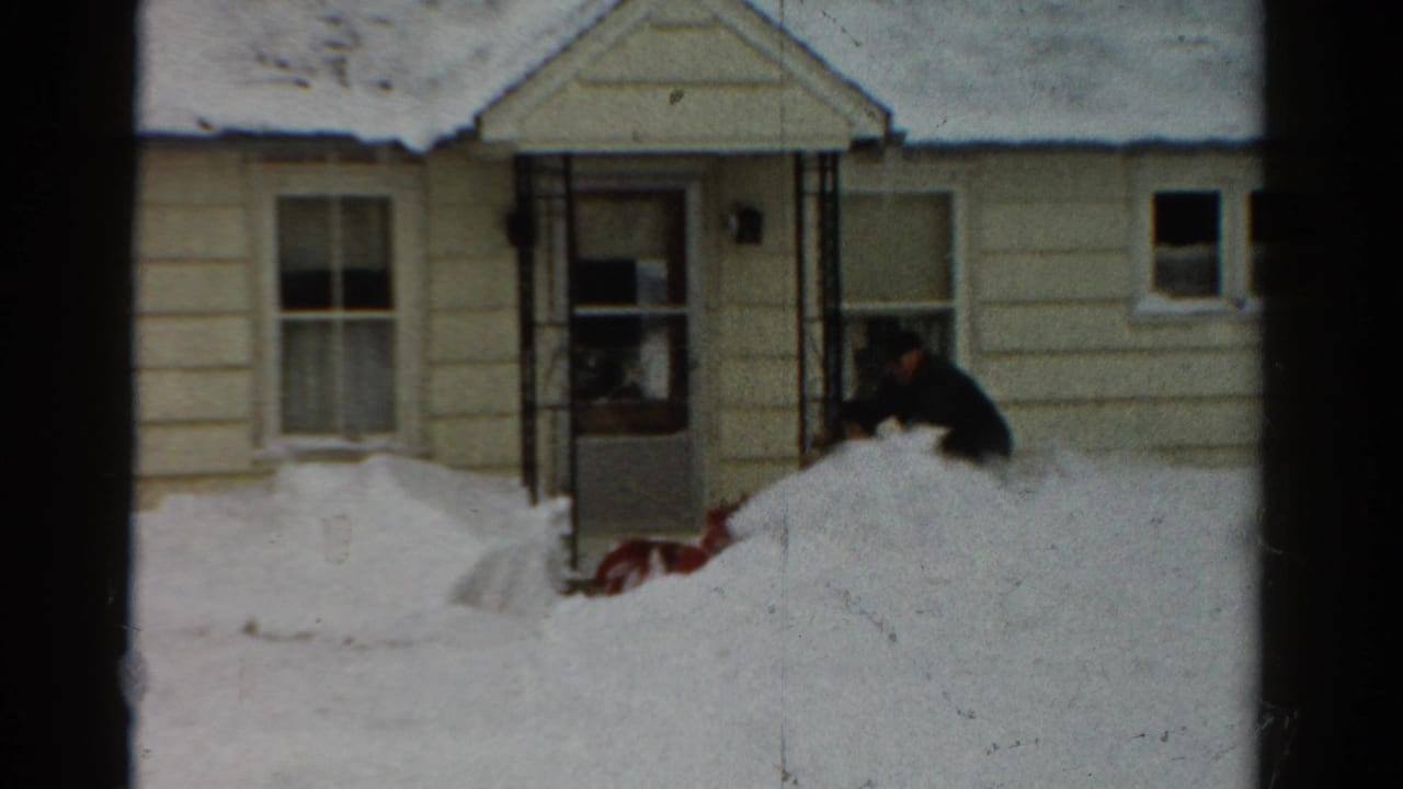In 1954 Toledo, a man works a red snow blower near the front entry of a snow-covered beige house.