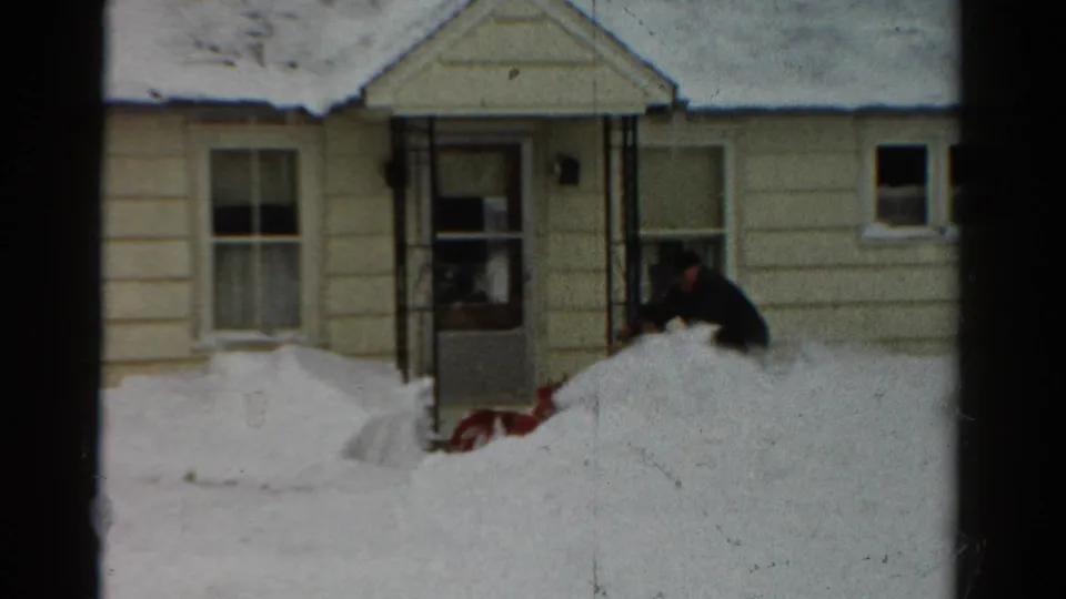 In 1954 Toledo, a man works a red snow blower near the front entry of a snow-covered beige house.