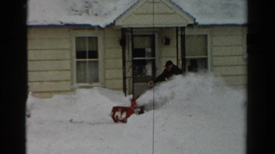 The man guides the red snow blower while snow sprays into a pile near a Toledo house entrance.