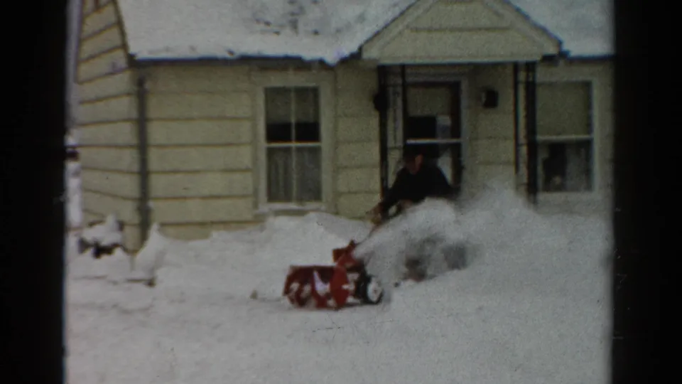 A fixed 8mm view shows the man pushing a red snow blower through snow outside the beige-sided Toledo home.