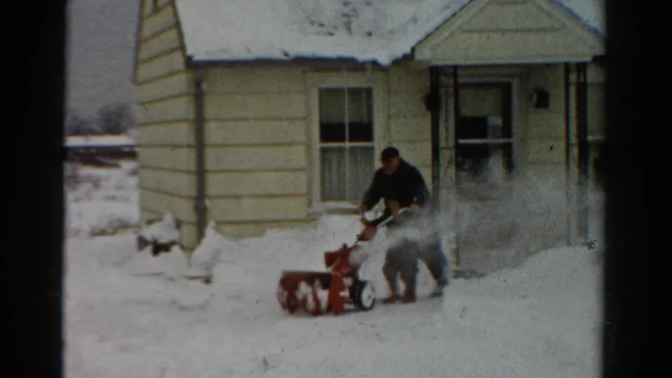 The man operates the snow blower near the house, with a broad plume of snow rising beside him in the 1954 winter scene.