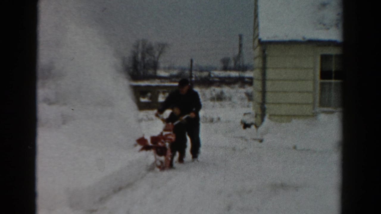In 1954 Toledo, Ohio home-movie footage, a man in dark winter clothing operates a red snowblower beside a light-colored house.