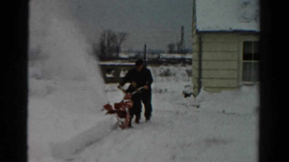 In 1954 Toledo, Ohio home-movie footage, a man in dark winter clothing operates a red snowblower beside a light-colored house.