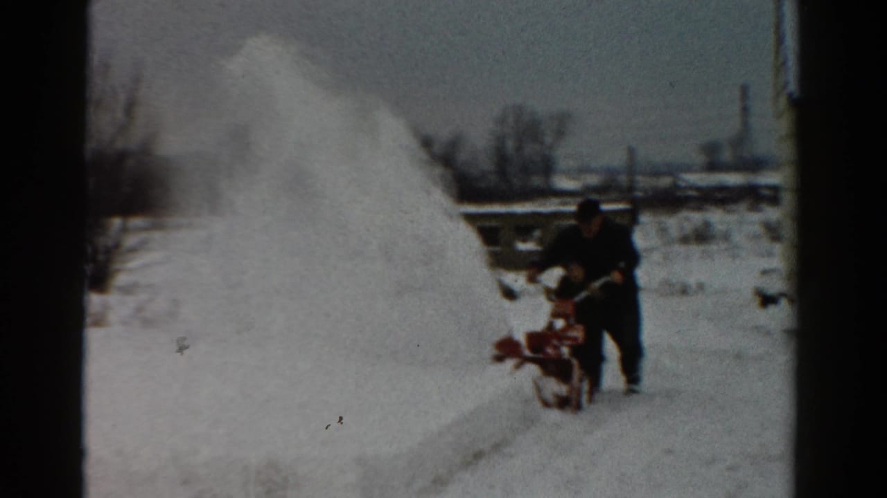 Man stands behind a red snowblower beside a high bank of snow in a yard.