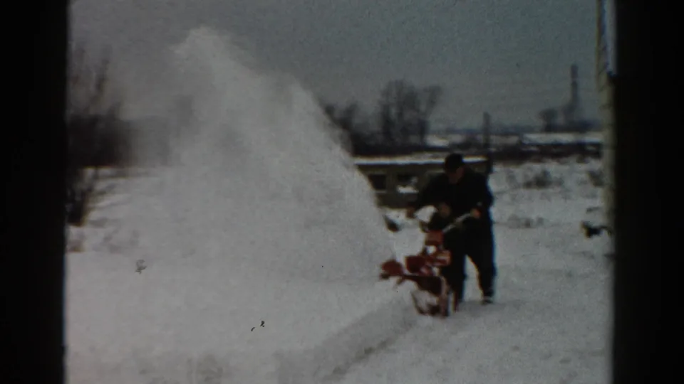 A man operates a red snowblower in a snowy Toledo yard, with a house wall, bare trees, and heavy snowbanks visible.