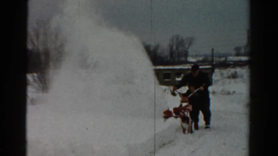 The 8mm frame shows a man in winter clothing pushing a red snowblower through deep snow in a residential Toledo setting.