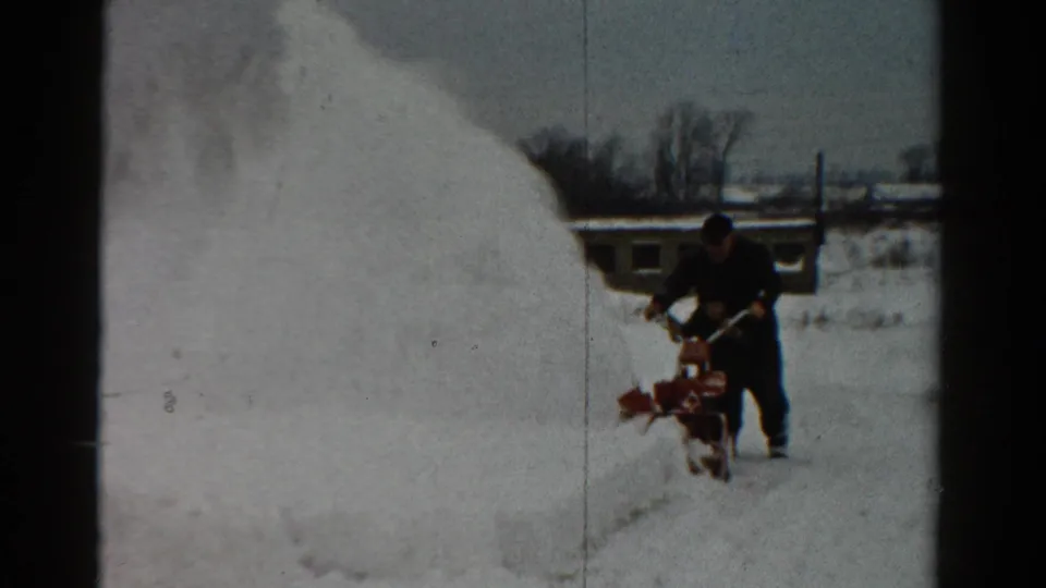 A man operates a red snowblower across a snow-covered yard in 1954 Toledo, with bare trees and a low structure in the background.