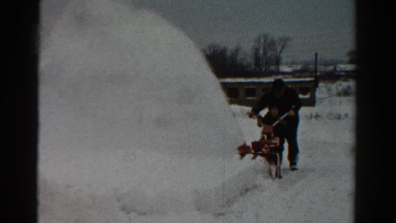 Man pushes a red snow blower through deep snow beside a large snowbank.