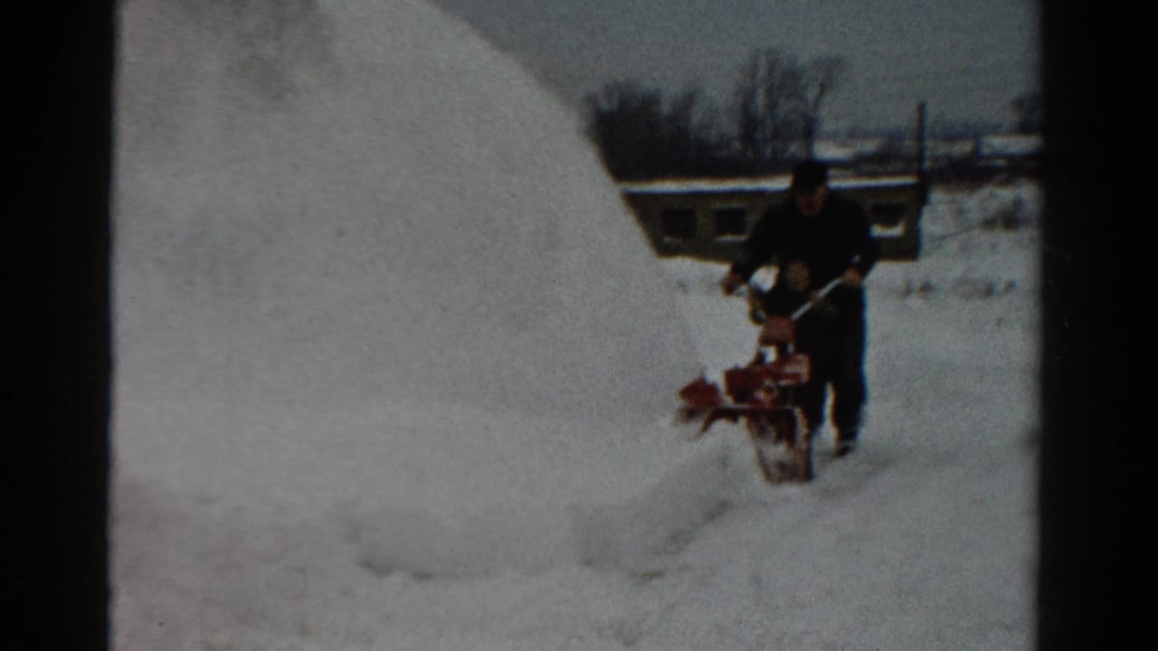 Man continues guiding a snow blower along a narrow cleared track in deep snow.