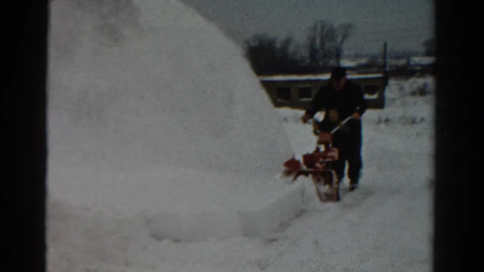 A lone man operates a red snow blower in a snowy Toledo yard, with bare trees and a low building behind him.
