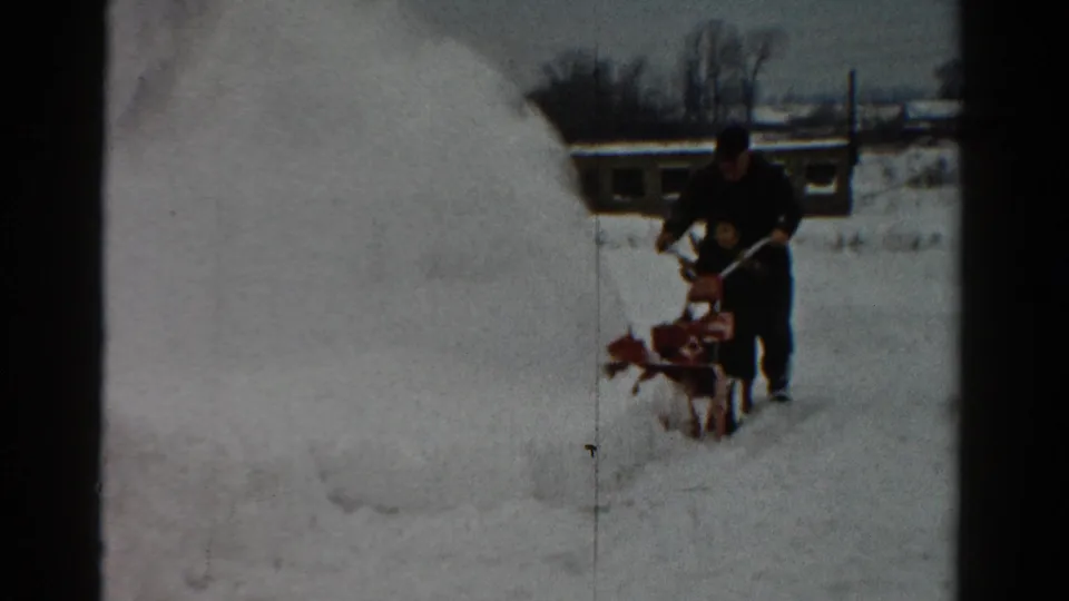 The home-movie frame shows a man in winter clothing pushing a red snow blower through deep 1954 Toledo snow.