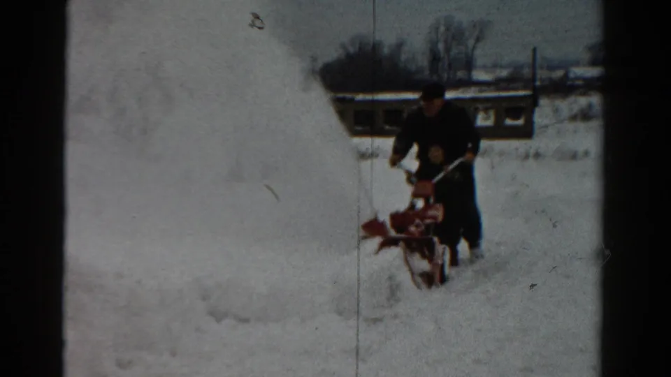 A man uses a walk-behind red snow blower near a high snowbank in a snowy Toledo residential scene.