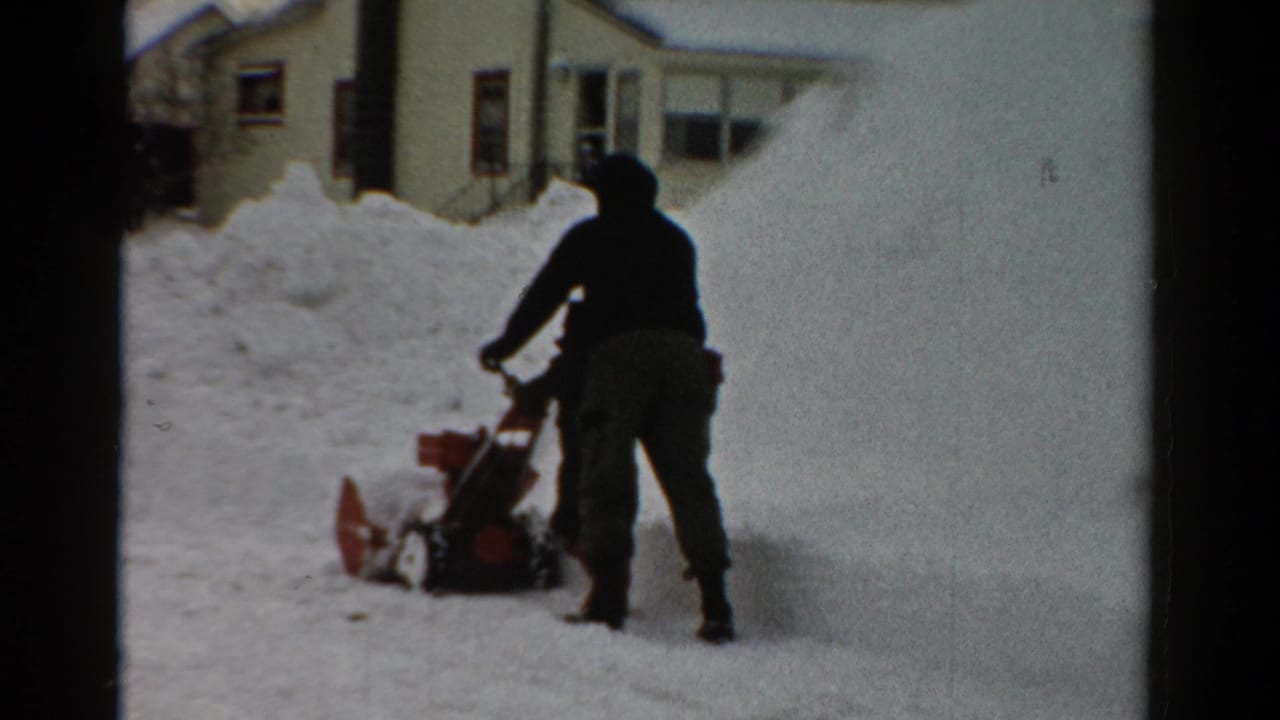 Hooded person pushing a red snow blower beside deep snowbanks in a residential yard.