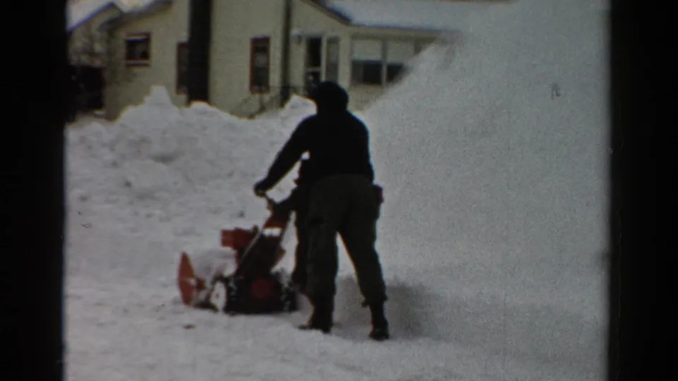 1954 Toledo 8mm frame showing a hooded winter figure operating a red snow blower near deep snow and houses.