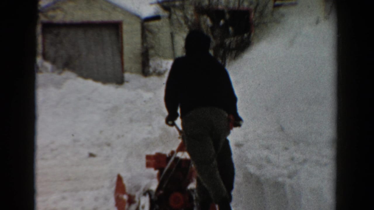 Hooded figure seen from behind pushing a snow blower toward a snowy garage area.