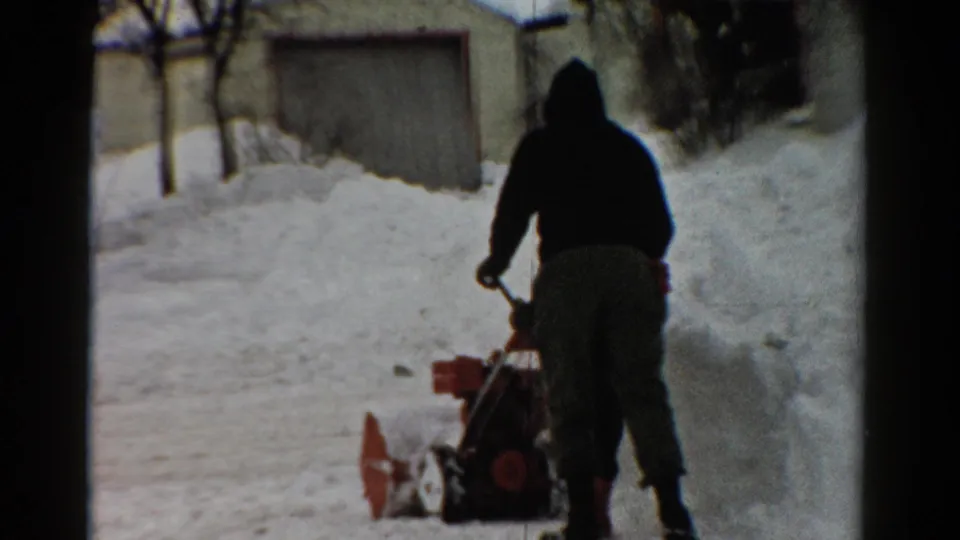 1950s 8mm frame from Toledo showing a winter-clad figure operating a snow blower in a snowy residential area.