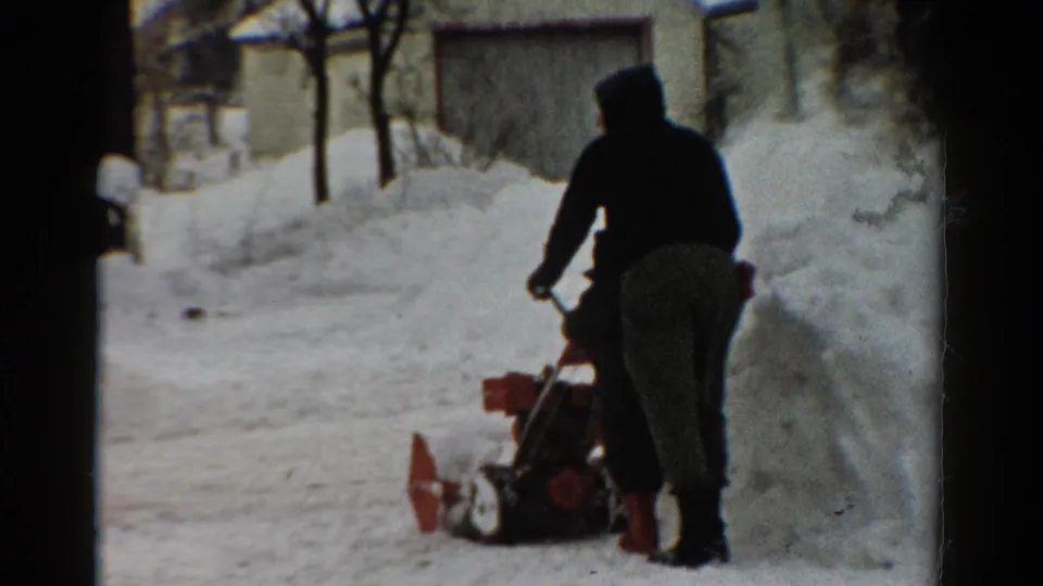 1954 Toledo home-movie frame of a hooded figure using a red snow blower beside snowbanks and winter trees.