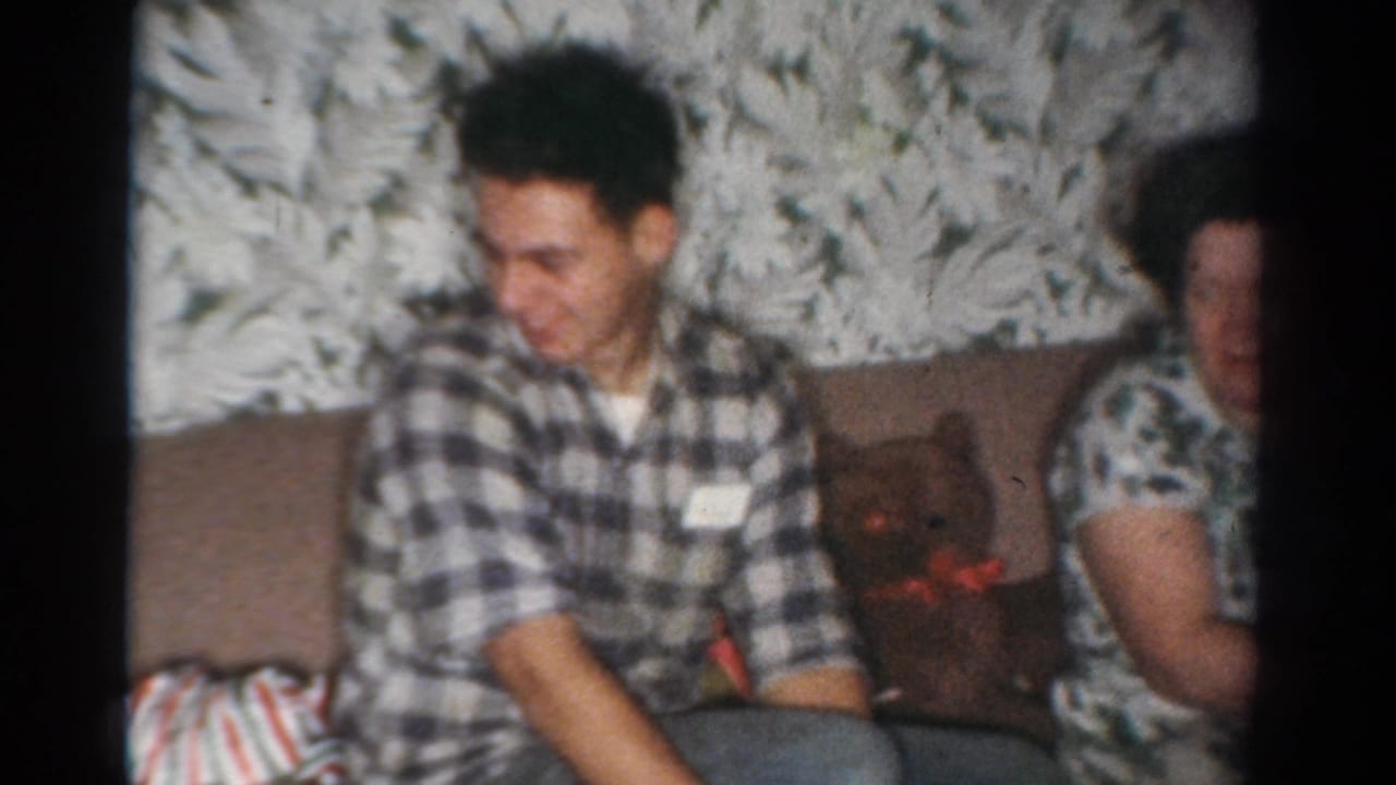 In a 1954 Toledo home-movie frame, a man in a checked shirt sits on a couch beside a woman with gifts and holiday decor nearby.