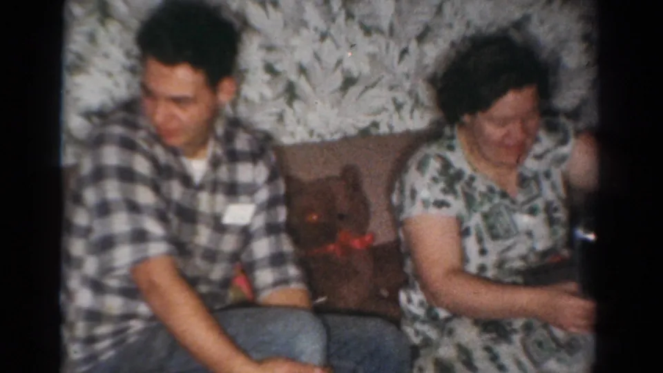 In the Toledo Christmas home movie, a man in a checked shirt and a woman in a patterned dress sit near a small holiday decoration.