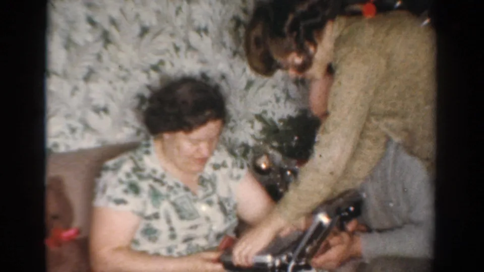 A woman in a patterned dress sits on the couch as another adult leans forward near a small Christmas tree and wrapped gifts.