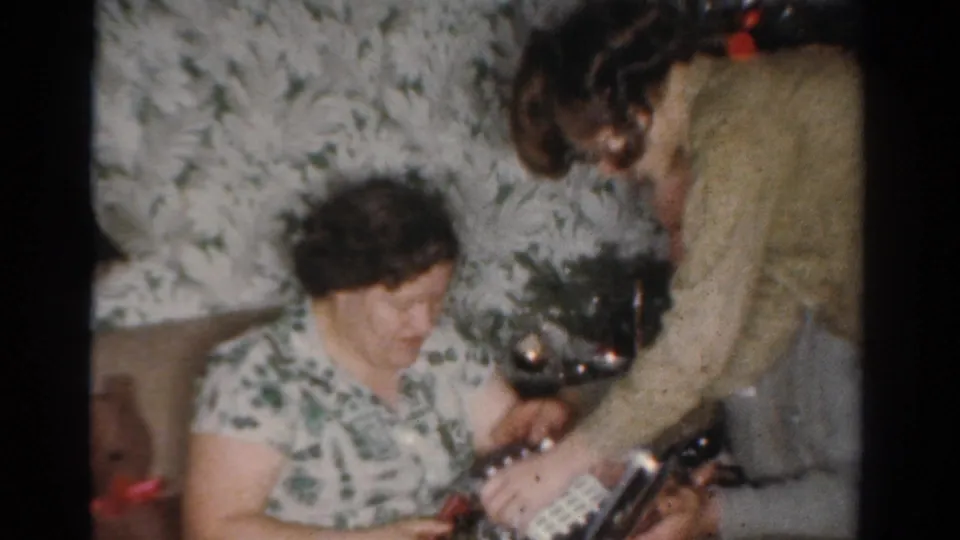 In the 1954 Toledo holiday scene, a seated woman smiles while another adult reaches toward a gift near a small decorated tree.